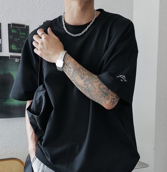 Person wearing a black oversized t-shirt with visible tattoos and a watch, standing against a neutral background.