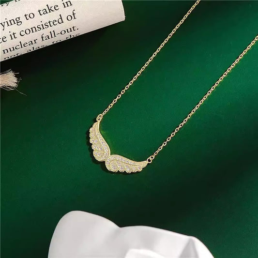 Gold angel wing necklace on a green surface with a white fabric and newspaper.