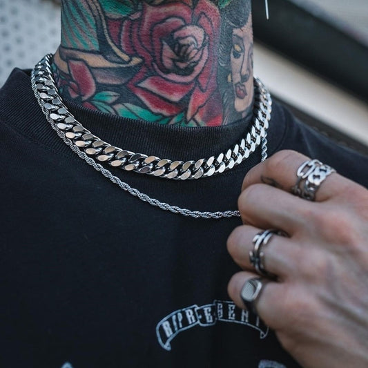 Person wearing multiple silver chains and rings with tattoos on neck and chest.