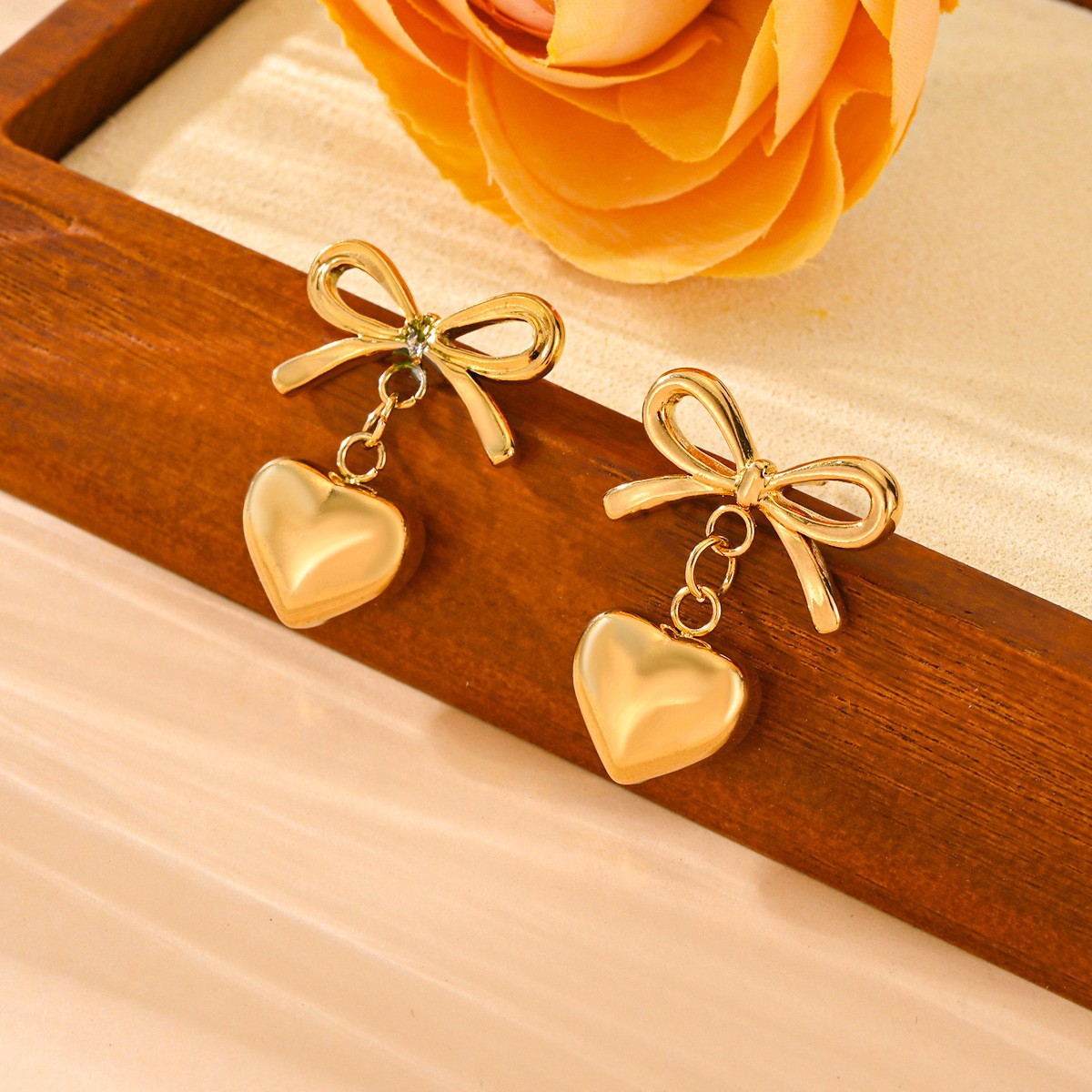 Gold bow and heart-shaped earrings on a wooden tray with flowers in the background