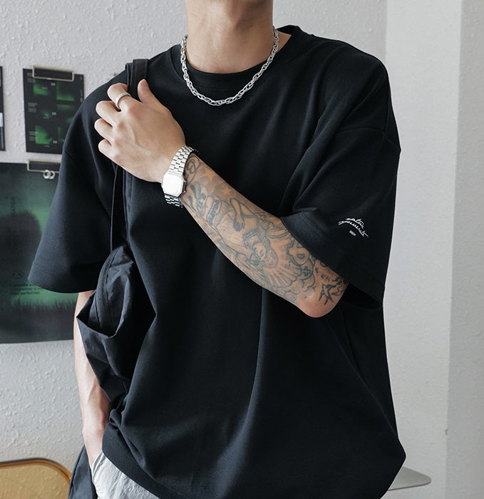 Person wearing a black oversized t-shirt with visible tattoos and a watch, standing against a neutral background.