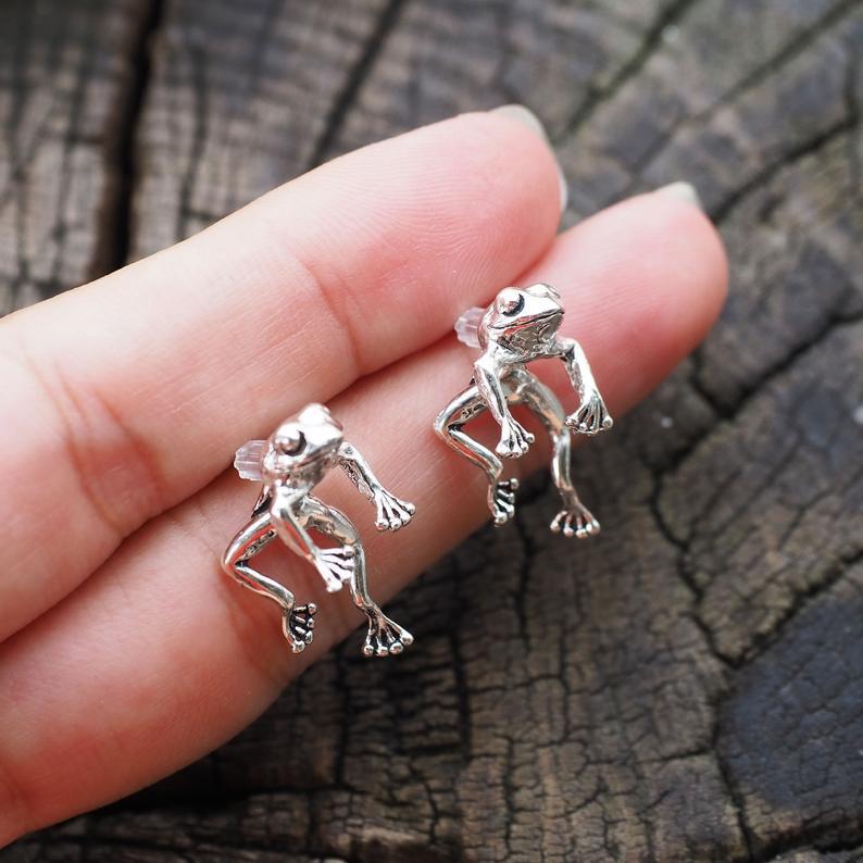 Silver frog earrings held between fingers against a wooden background