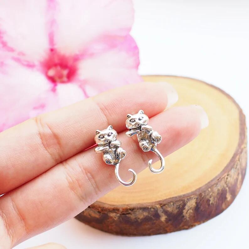Silver cat-shaped earrings held between fingers with a pink flower in the background