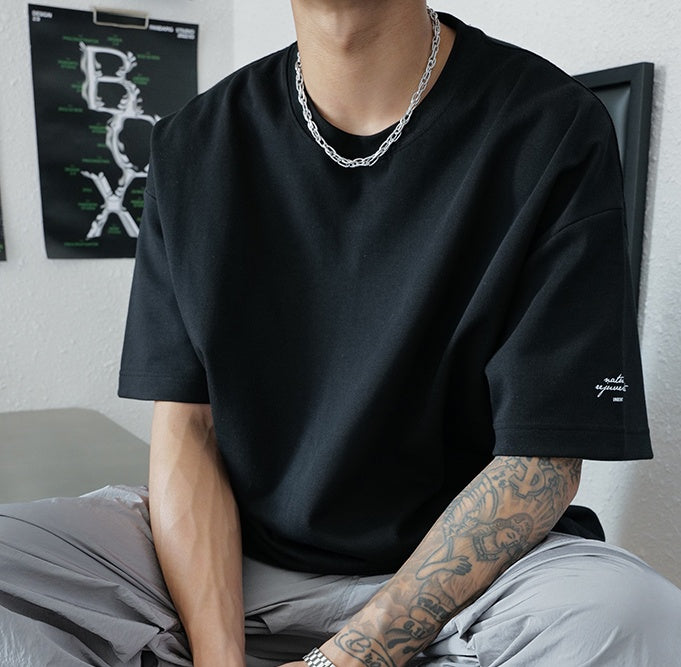 Person wearing a black t-shirt with a brand logo, sitting on a bed.