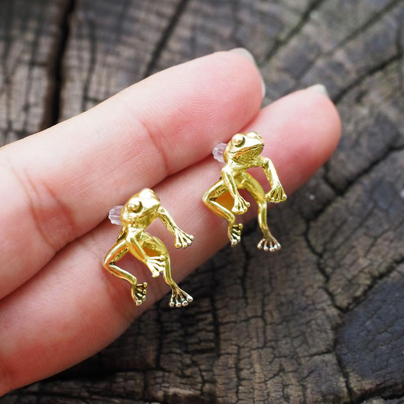 Gold frog-shaped earrings held between fingers against a wooden background