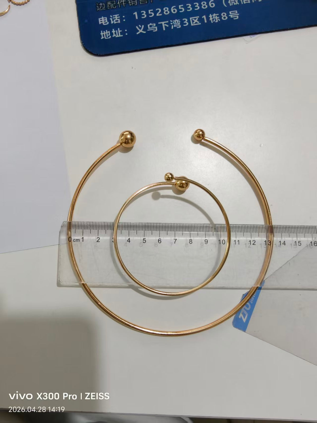 A pair of gold-plated hoop earrings with a small bead detail at the center, shown on a white surface with a clear ruler for scale.