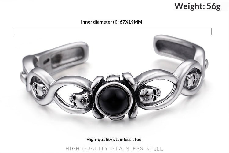 A silver-toned stainless steel cuff bracelet with skull motifs and a central black oval stone, shown on a white background with text indicating inner diameter and weight.